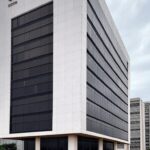 Edifício da Caesb em Brasília com documentos de concurso sob céu nublado, representando frustração de candidatos no DF.