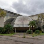 Fachada da Câmara Legislativa do DF em Brasília, com pódio vazio e prédios de seguros ao fundo, simbolizando homenagem criticada por ignorar crises.