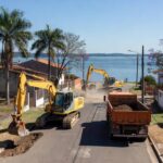 Obras de infraestrutura no Paranoá, com equipamentos de construção em rua residencial e Lago Paranoá ao fundo.